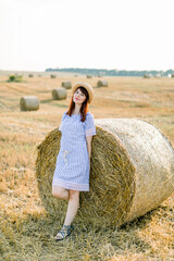 Naklejka na ściany i meble Countryside landscape of farm wheat field at summer sunset. Pretty young happy Caucasian woman in straw hat and blue striped dress posing near the hay bale and enjoying warm evening and vacations