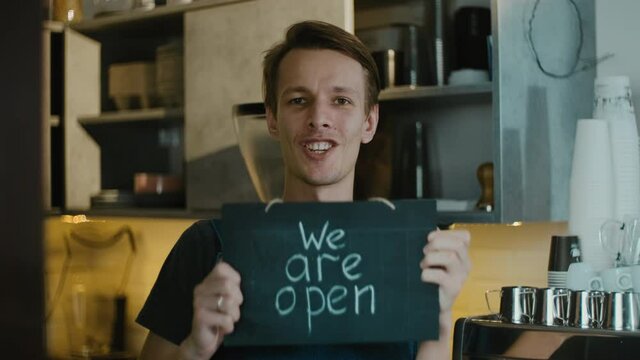 Excited and happy coffee shop owner invites new guests holding board with sign we are open after finishing quarantine and lockdown is lifted. Support local business
