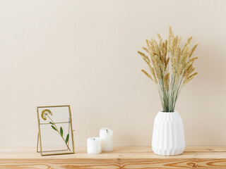 Composition of white ceramic vase with bouquet of dry spikelets, golden photo frame with dried flower, candles on wooden table on pastel beige background. Stylish home decor. Modern interior design.