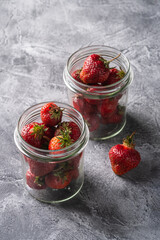 Fresh ripe strawberry fruits in two glass jars, summer vitamin berries on grey stone background, angle view