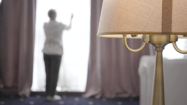 Close-up of floor lamp standing in hotel room with blurred Caucasian maid opening curtains at the background. Wide shot of professional woman adjusting drape in accommodations. Tourism, travelling.