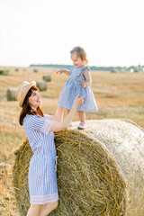 Naklejka na ściany i meble Happy mother and little daughter, wearing similar striped dresses, having fun in summer field with golden hay bales. Child on a haystack. Countryside, happy childhood