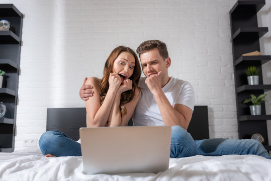 Emotional Couple Watching Scary Movie On Laptop In Bedroom