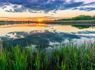 Scenic view at beautiful spring sunset with reflection on a shiny lake with green reeds, bushes, grass, golden sun rays, water ,deep blue cloudy sky and glow on a background, spring evening landscape