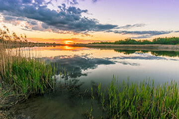 Scenic view at beautiful spring sunset with reflection on a shiny lake with green reeds, bushes, grass, golden sun rays, water ,deep blue cloudy sky and glow on a background, spring evening landscape