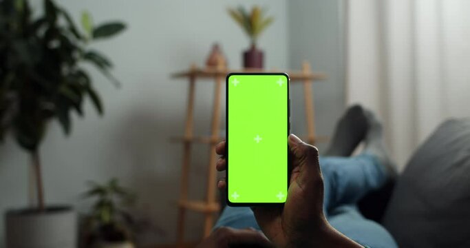 Afro american man holding mobile phone in one hand and looking at mock up screen with trecking markers. Guy lying on sofa and watching video while using smartphone. Concept of chroma key.