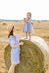 Naklejka na ściany i meble Happy mother and little daughter, wearing similar striped dresses, having fun in summer field with golden hay bales. Child on a haystack. Countryside, happy childhood