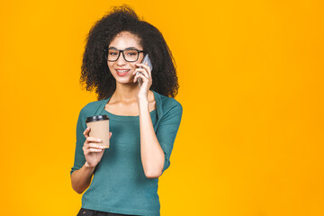 Happy young african american woman casually dressed standing isolated over yellow background, talking on mobile phone, holding takeaway cup of coffee or tea.