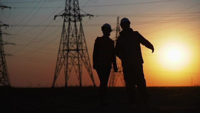 team of engineers in protective helmets discuss and gesticulate with hands while discussing construction of new power line for supplying electric power to city at sunset, silhouette