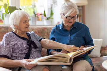 Senior woman and her adult daughter looking at photo album together on couch in living room
