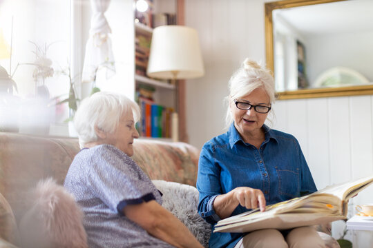 Senior woman and her adult daughter looking at photo album together on couch in living room
