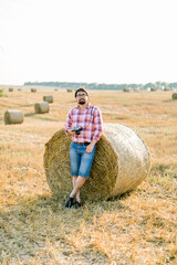 Naklejka na ściany i meble Handsome bearded brunette man, wearing checkered shirt and jeans leaning on a hay bale in goldern wheat field, posing, holding retro camera in hands, and enjoying warm sunset in summer day