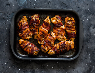 Roasted bbq chicken breast wrapped in prosciutto on a baking pan and grilled bread on a dark background, top view. Delicious tapas, snack, appetizer