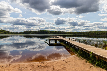 Fototapeta na wymiar Landscape with a swamp lake and with a wooden footbridge. Beautiful clouds and the surrounding forest were reflected in dark water. Latvia. Lake Ratnieku