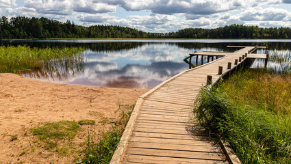 Fototapeta premium Landscape with a swamp lake and with a wooden footbridge. Beautiful clouds and the surrounding forest were reflected in dark water. Latvia. Lake Ratnieku
