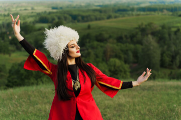 Georgian girl in white papakha and red national dress dances national dance on the green hills of...