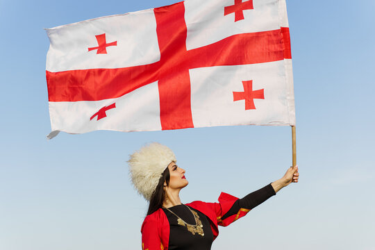 Georgian girl with national flag of Georgia in hands on blue sky background. Georgian culture lifestyle. Woman in papakha and red dress