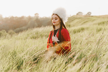 Georgian girl with closed eyes in white papakha and red national dress seats on the green grass and...