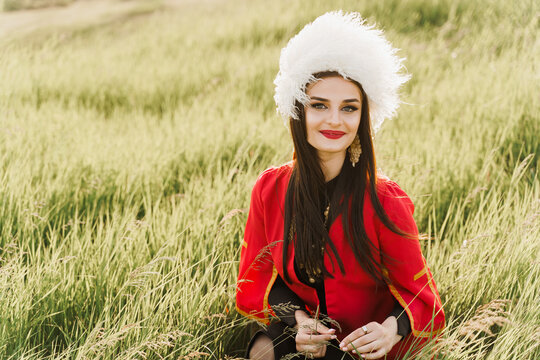 Georgian girl in white papakha and red national dress seats on the green grass and smiles into camera. Georgian culture lifestyle.