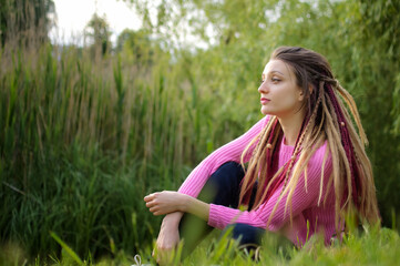 Outdoors female portrait of a girl with dreadlocks wearing pink sweater in a city park during a sunset, be free concept.