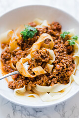 A fork is full of freshly homemade pappardelle pasta noodles in a meat bolognese sauce. This is Italian home cooking. The spaghetti bolognese is served in a white bowl and garnished with parsley.