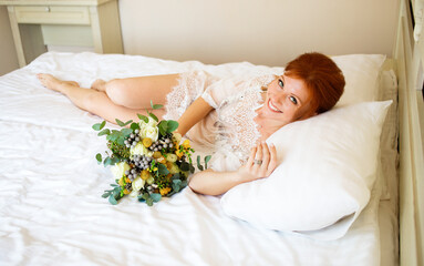 Obraz na płótnie Canvas The beautiful red-haired bride in a transparent peignoir lies on the bed with a bouquet in her hands. Boudoir morning of the bride. Top view.