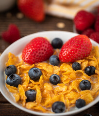 Corn flakes with blueberries and raspberries in white bowl in dark wooden desk.
