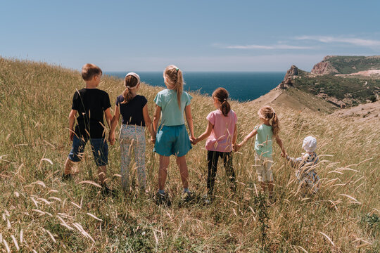 six children of different ages back to the camera, girls and boys hold hands walking across the field in the grass against the sea and sky. It's a sunny summer day.