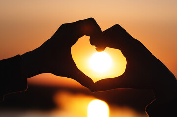 silhouette of female hands forms a heart on a sunny sunset sky. love and gratitude. beautiful view on the beach