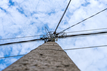 An electric pole with wires rising up towards a blue sky with clouds. At the top of the pole,...