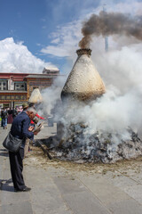 Smoking sangkang incense burner on Barkhor square with a man pilgrim wearing shaggy cloak in a bow with hat off