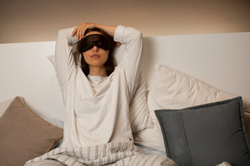 Young attractive caucasian girl in white pajamas and sleep mask in bed with pillow getting ready for bed. Rest at home. Healthy night sleep. Сozy interior, stylish lighting.
