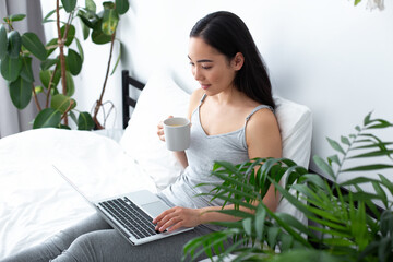 Merry young woman relaxing with notebook and coffee in bedroom