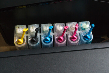Dirty ink cartridges connected to a black printer. Six jars of different colors stand in a row.