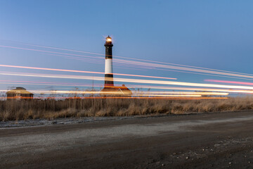 Car lights streaking by a lighthouse has they drive down a dirt road. Fire Island, New York