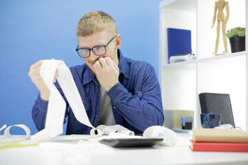 Fototapeta premium Frustrated caucasian male in glasses, dressed in casuals holds a long receipt in hands, considers the amount of debt and feels anxious, bites finger nails in puzzlement, looks nervously at invoices