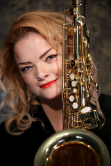 Obraz premium Portrait of a charming blonde with a saxophone in her hands. Beautiful girl in a black evening dress. Young woman with her hair and bright red lips. Face close up. Saxophonist lady