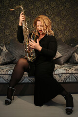 Naklejka premium Portrait of a charming blonde with a saxophone in her hands. Beautiful girl in a black evening dress. Young woman with her hair and bright red lips. Face close up. Saxophonist lady
