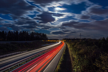 Aerial view of highway at night. Rear red and front white light trail visible on road. Long exposure photography. Full moon and illuminated clouds on the sky