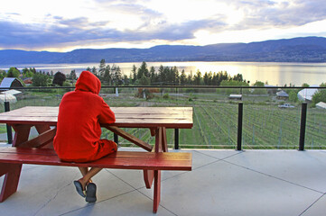 Social distancing during Covid-19 pandemic. The view on man in red hoodie sitting by the wooden picnic table on the patio with the view on Okanagan vineyards and lake. Kelowna, BC.
