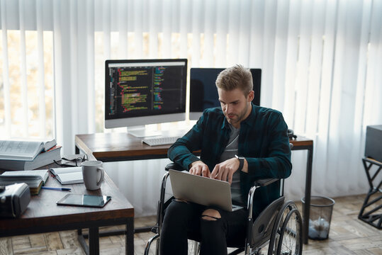 Multitasking. Portrait of young male web developer in a wheelchair writing program code on his laptop at his workplace in the office. Multiple computer screens with program code