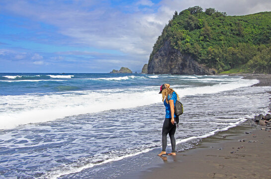 Pololu valley, Big Island, Hawaii. The detail on the female / girl hiker with backpack standing bare foot on the black sand beach, watching the waves in the ocean. The green cliff in the bacgrkound. - Powered by Adobe