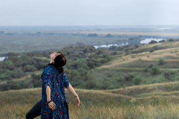 Dark-haired beautiful flexible girl in a black top and leggings and a turquoise tunic dancing in nature. Beautiful landscape, river in the background.