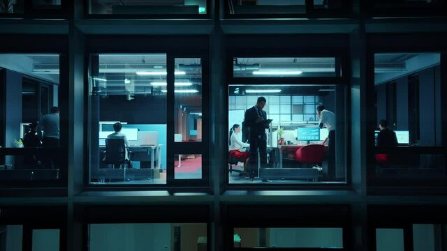 Footage from Outside the Window: Businessmen and Businesswomen Working in the Office. Managers and Specialists Doing Financial Business in the Evening. Employees Work on Computers and Delegate Tasks.