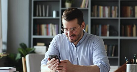 Excited young businessman in eyeglasses reading message with good news on smartphone. Happy euphoric male user looking at cellphone screen, celebrating online win, mobile victory success concept.