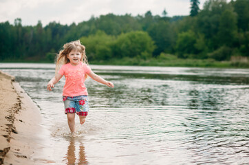Little beautiful girl runs along the riverbank looking at the camera and laughing, a lot of spray and sunlight