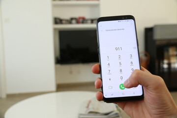 A hand holding a generic new mobile phone which displays the emergency number 911 on the touch screen. A concept about calling for help from inside a house. An image on an interior background.