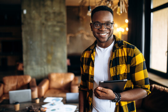 Portrait of attractive smiling African American guy, student or employee, in yellow checkered shirt standing in office with tablet in hand in a positive mood