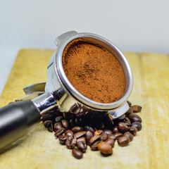 Fresh grounded coffee in espresso machine porta filter surrounded by coffee beans on wooden plate.
