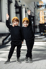 Two cheerful embraced twin boys in casual outfit posing with hands up in the street.
